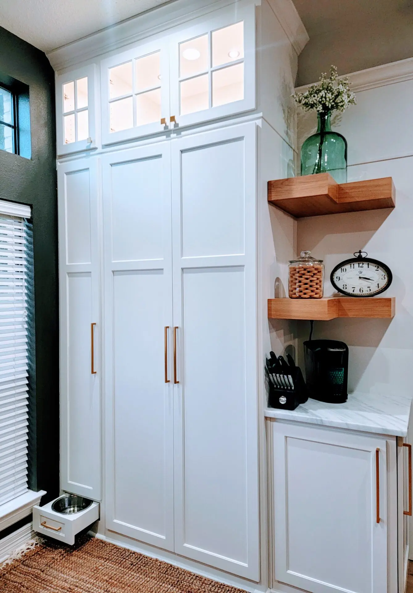 Exceptional custom white built-in cabinetry with upper glass panels, adjacent floating shelves holding decorative items and a clock, with a pet feeding bowl at the bottom.