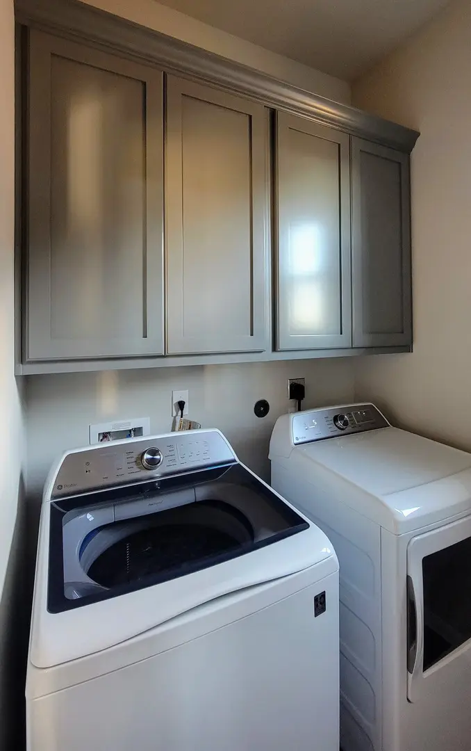 A modern laundry room with a top-loading washing machine and a front-loading dryer under grey cabinets, enhanced by exceptional custom furnishing.