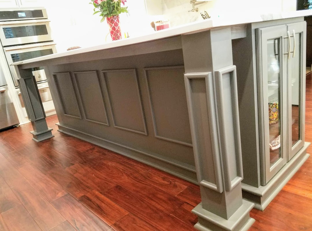 A kitchen island with gray paneling and glass cabinet doors on polished wooden flooring, featuring exceptional custom furnishing.