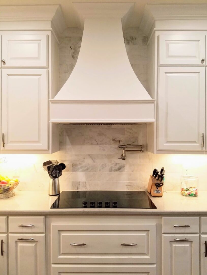 A modern kitchen with exceptional custom furnishing, white cabinetry, marble backsplash, and a stainless steel cooktop under a range hood.