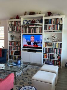 A well-organized living room with exceptional custom furnishing, a large bookshelf, a television broadcasting a news channel, and comfortable furniture.