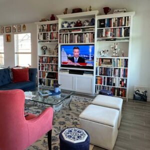 Cozy living room with exceptional custom furnishing, bookshelves, a television broadcasting news, and comfortable seating.