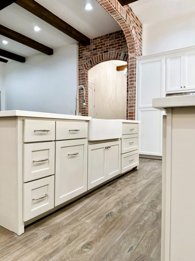 Modern kitchen interior with white cabinets, stainless steel handles, wood flooring, and a brick archway featuring exceptional custom furnishing.