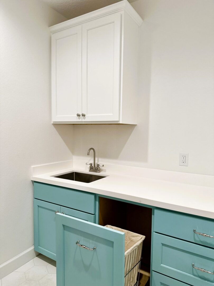 A minimalist laundry room with white walls, teal cabinetry, and a stainless steel sink, enhanced by exceptional custom furnishing.