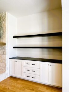 Built-in wall shelving unit with white cabinets and dark shelves against a light-colored wall, situated in a corner with a wooden floor.