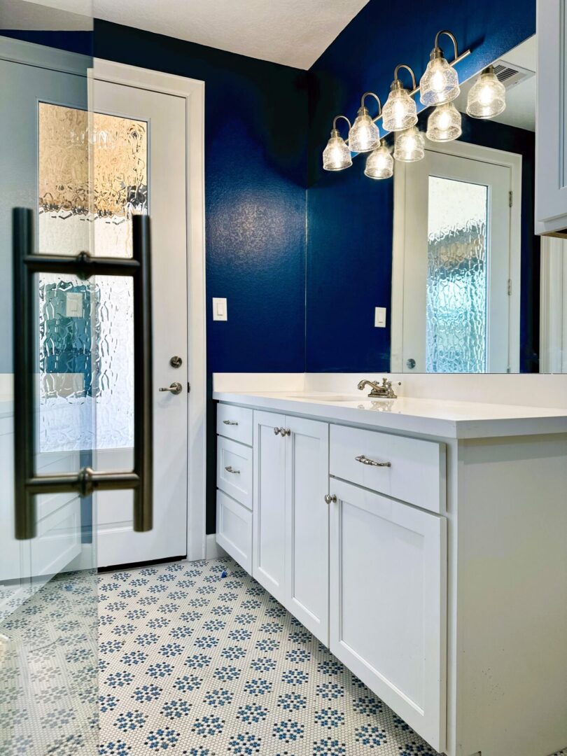 A modern bathroom featuring exceptional custom furnishing, a white vanity, patterned floor tiles, and a bold blue wall with multiple hanging light fixtures.