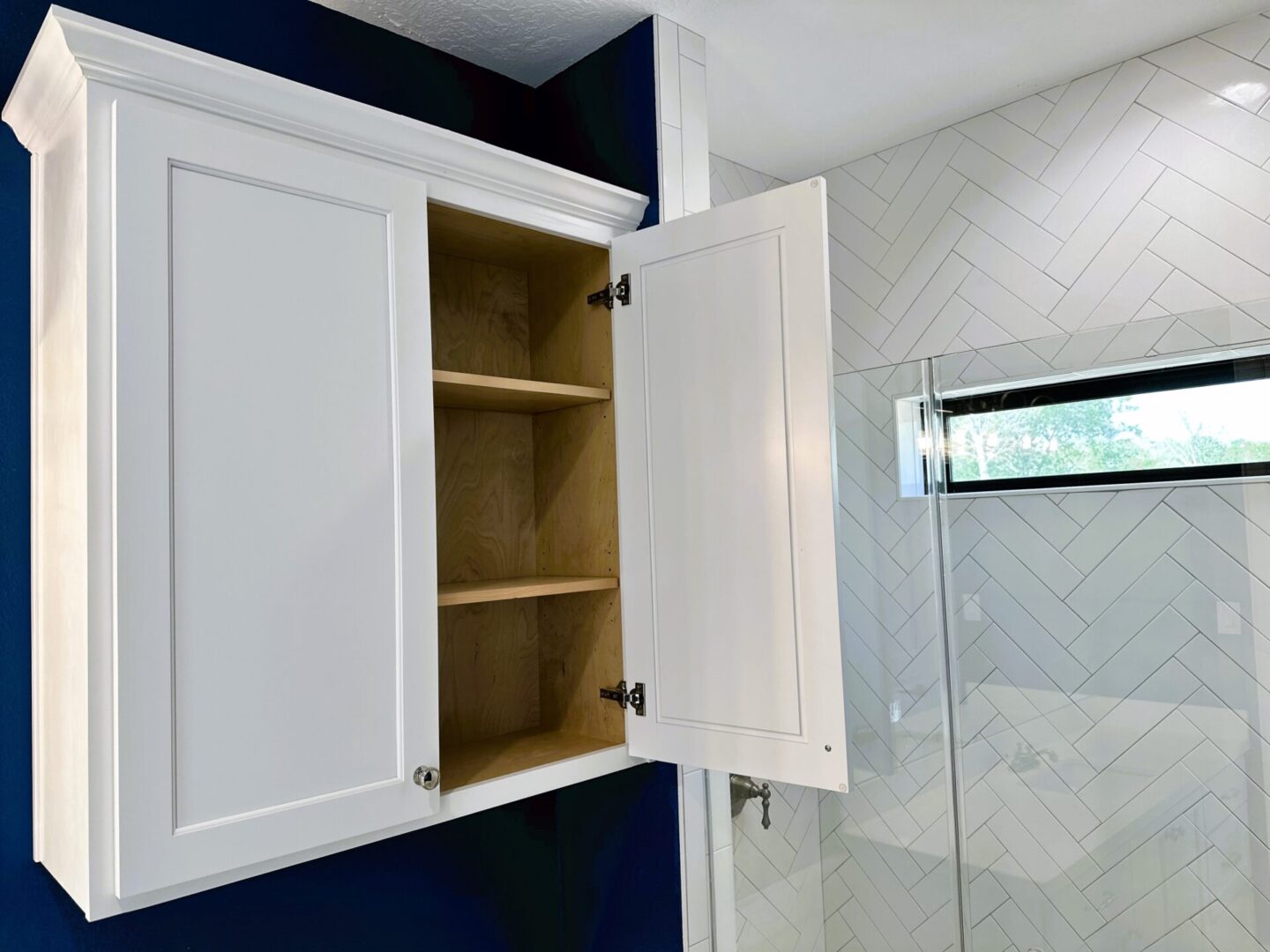 A white bathroom cabinet with its doors open, mounted on a wall with herringbone tile pattern, is an exceptional custom furnishing.