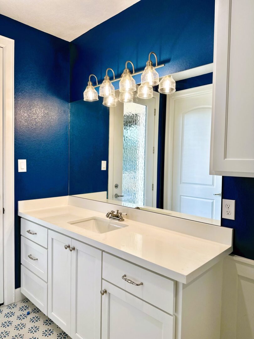 A brightly-lit bathroom with a white vanity, exceptional custom furnishing, a blue wall, and a mirror with glass light fixtures.