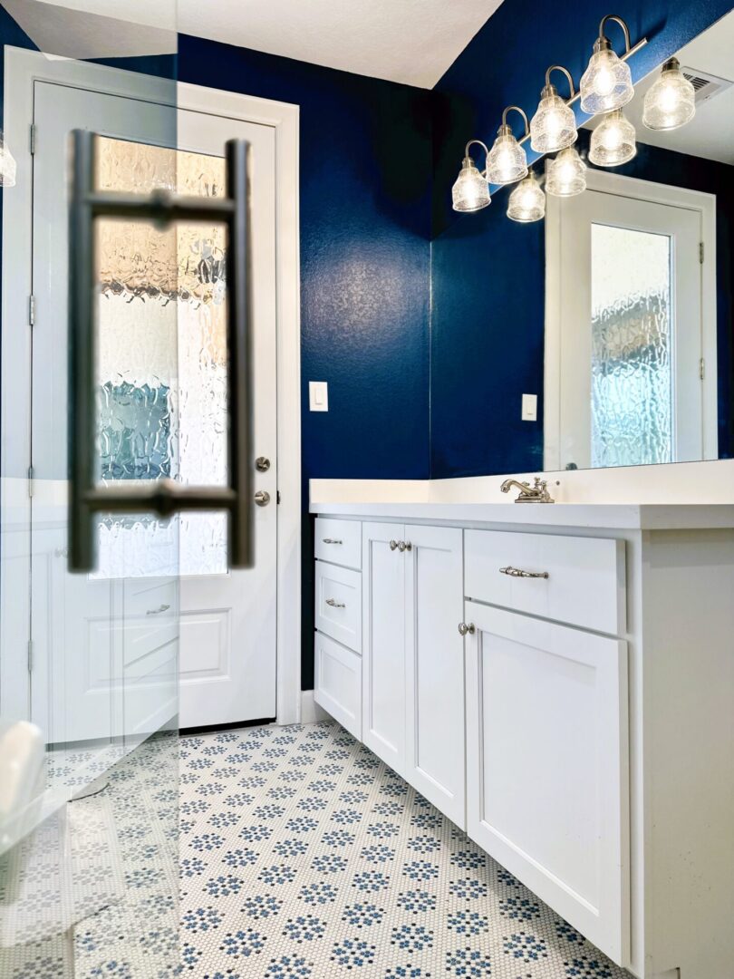 A modern bathroom with white cabinetry, patterned floor tiles, and a dark blue accent wall, featuring a glass-paned door partially open and an exceptional custom furnishing vanity mirror.