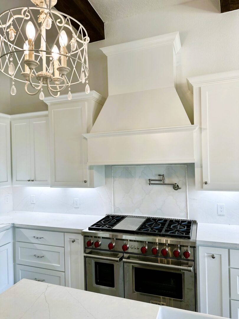 A modern kitchen featuring exceptional custom furnishing with white cabinetry, stainless steel double oven range, and a white marble backsplash under a large white range hood with a chandelier above.