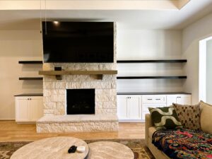 A modern living room featuring a stone fireplace with a mounted television above it, surrounded by floating shelves and white cabinetry.