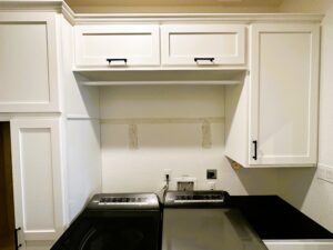 A kitchen with white cabinets where a range hood has been removed, leaving exposed walls and wiring.