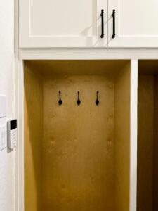 A wooden nook with three black hooks mounted on the back wall, situated beneath white cabinets.