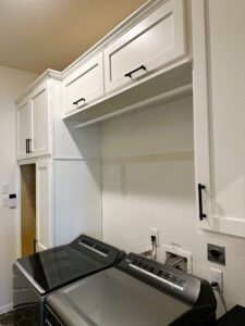 A laundry room with white cabinetry missing a section of backsplash over a washing machine.