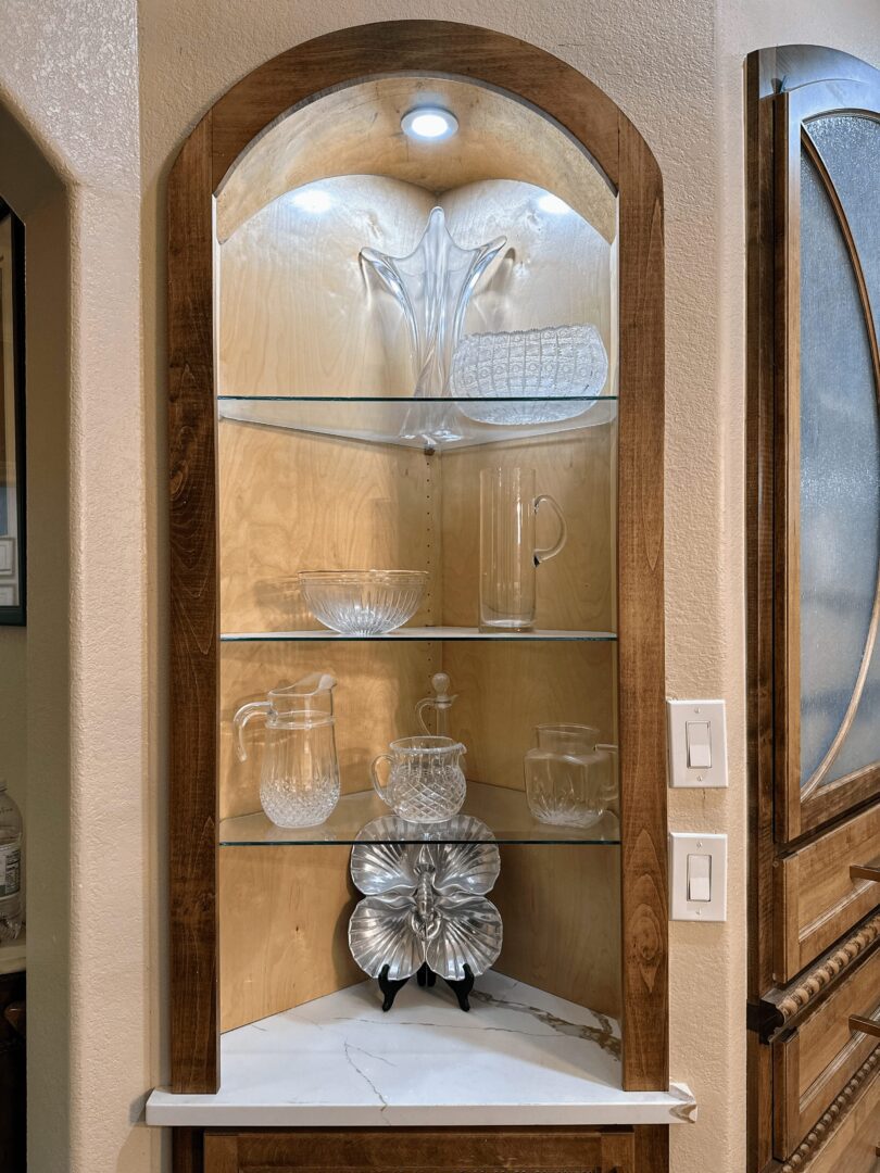 A wooden arched display cabinet with glass shelves showcasing various glassware and a decorative metallic piece on the lower shelf, illuminated by an overhead light.