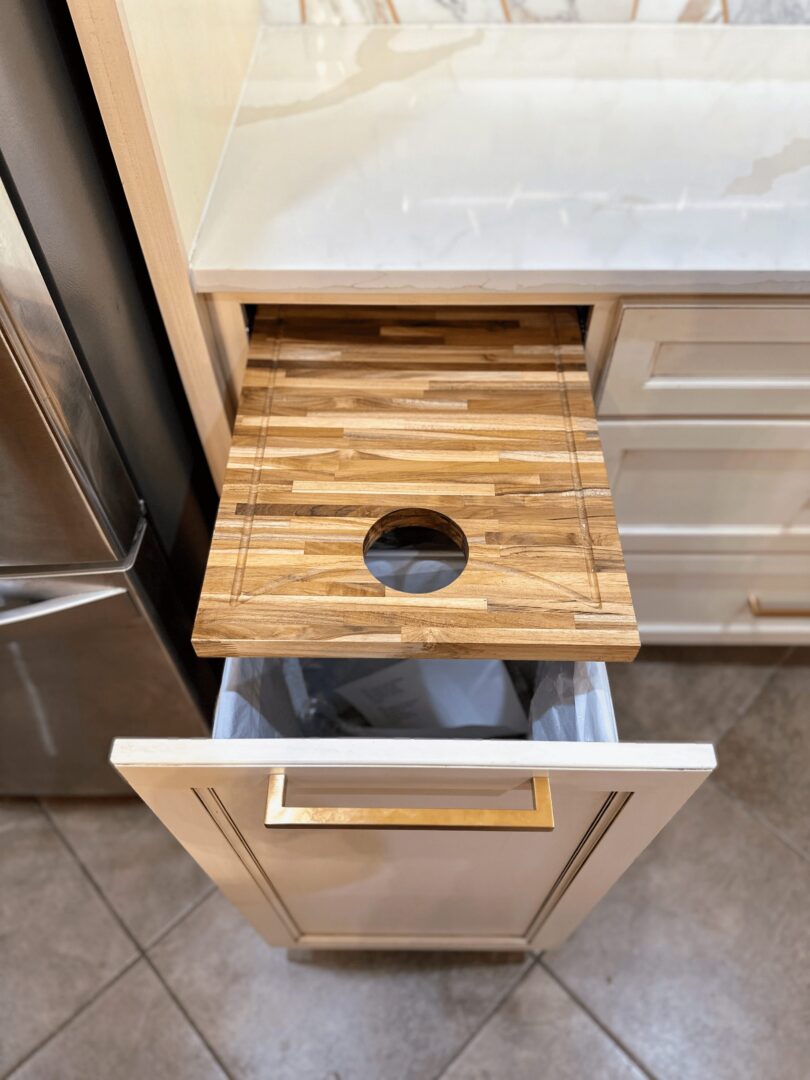 A kitchen drawer with a built-in cutting board featuring a hole for waste disposal directly above a trash bin.