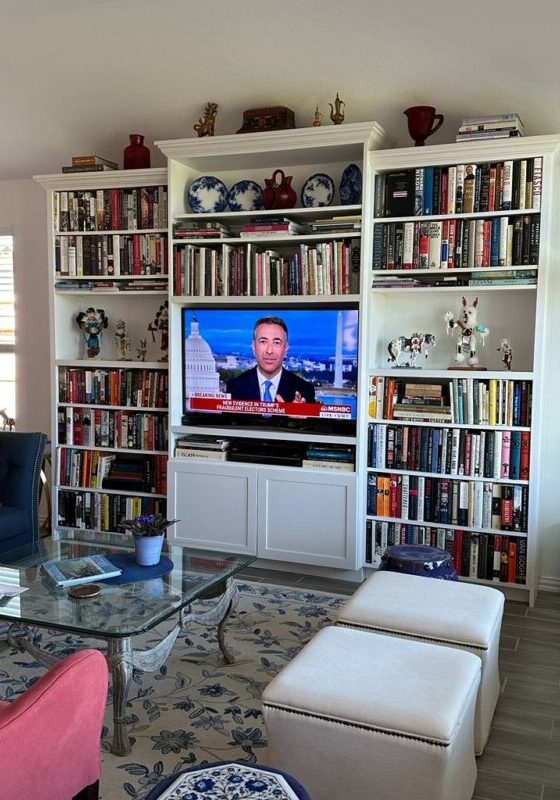 A well-organized living room featuring exceptional custom furnishing, with a large bookshelf surrounding a television broadcasting a news channel.