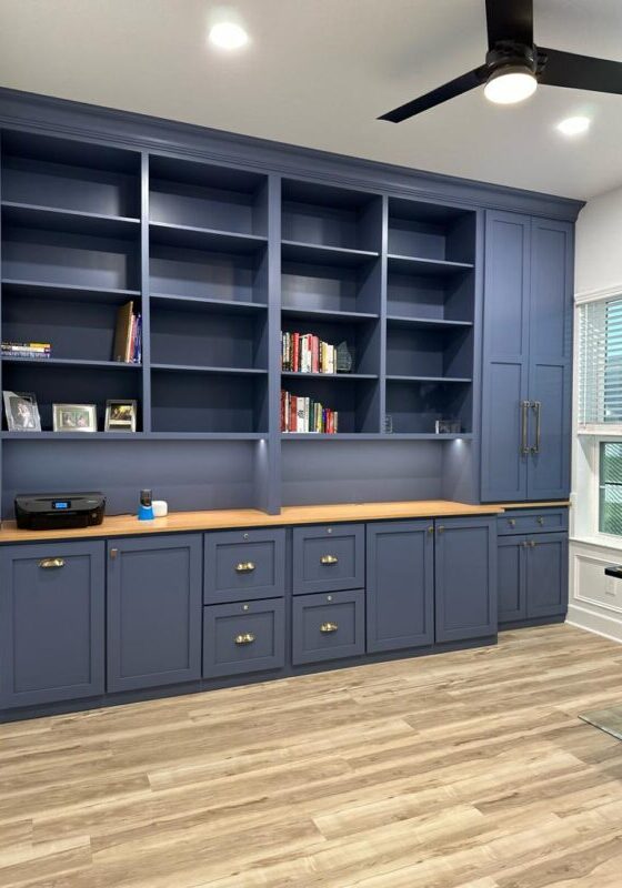 A modern home office with exceptional custom furnishing, featuring a blue built-in bookcase and cabinetry, a wooden floor, and a black ceiling fan.