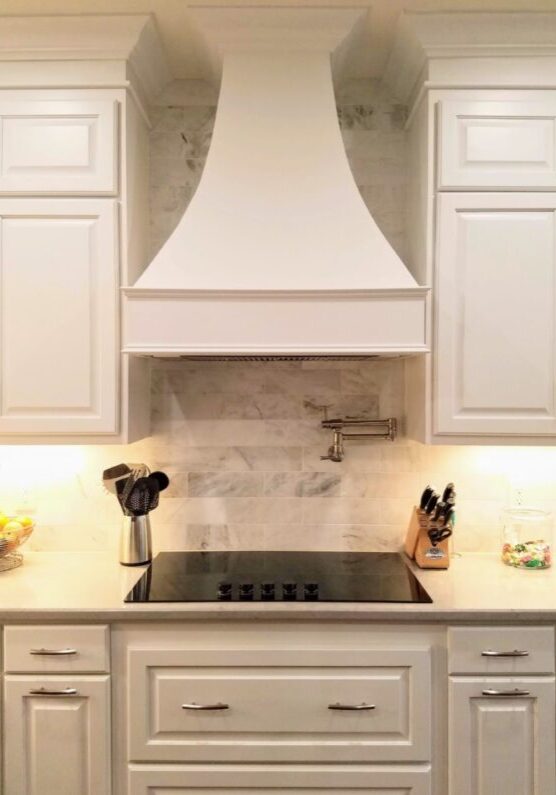 A modern kitchen with white cabinets, a marble backsplash, and a central cooktop under a white range hood features exceptional custom furnishing.